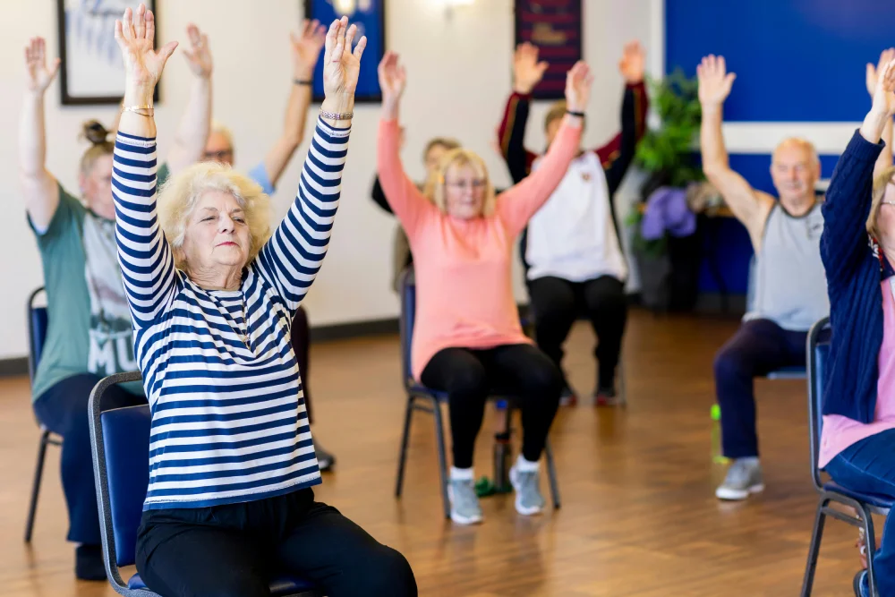 Elderly individuals participate in a group chair exercise class, promoting physical activity, mobility, and wellness in a supportive community setting.