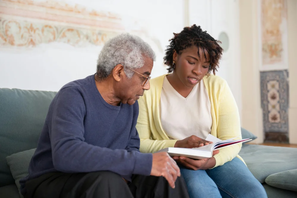 A young caregiver attentively reads a book with an elderly man on a cozy couch, fostering companionship and mental stimulation through shared activities.