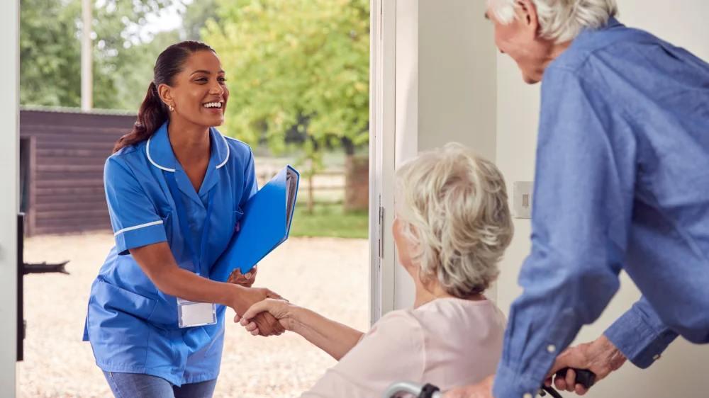 A caregiver in blue scrubs shakes hands with an elderly woman at the doorway while a man stands beside her.