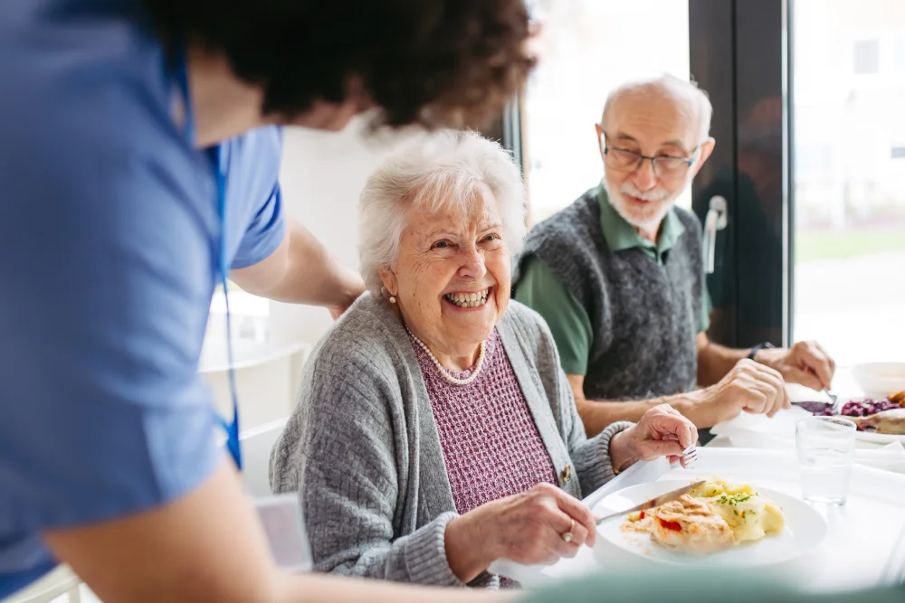 A smiling elderly woman eats lunch with a caregiver and another senior at a bright table.
