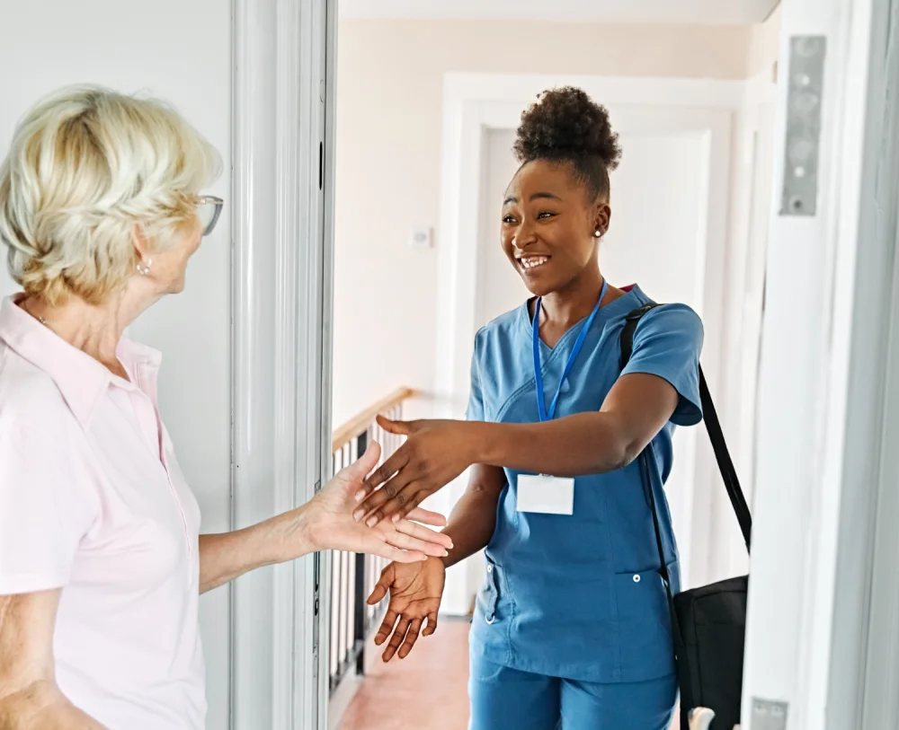 Caregiver in blue scrubs greets an elderly woman with a handshake in a home doorway.